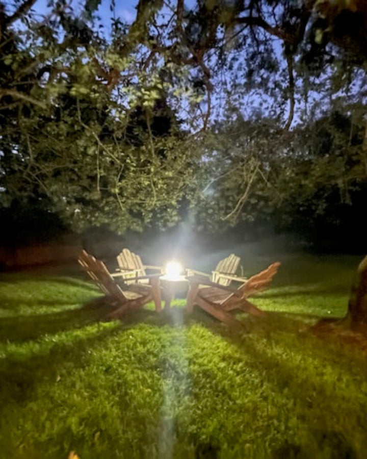 Adirondack chairs under a tree in a large backyard at night with a HAVEN solar portable lantern illuminating the area.