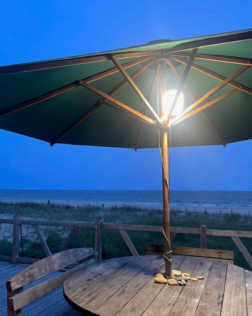 HAVEN portable solar lantern hanging from a patio umbrella on a wooden table with a beach view in the background.