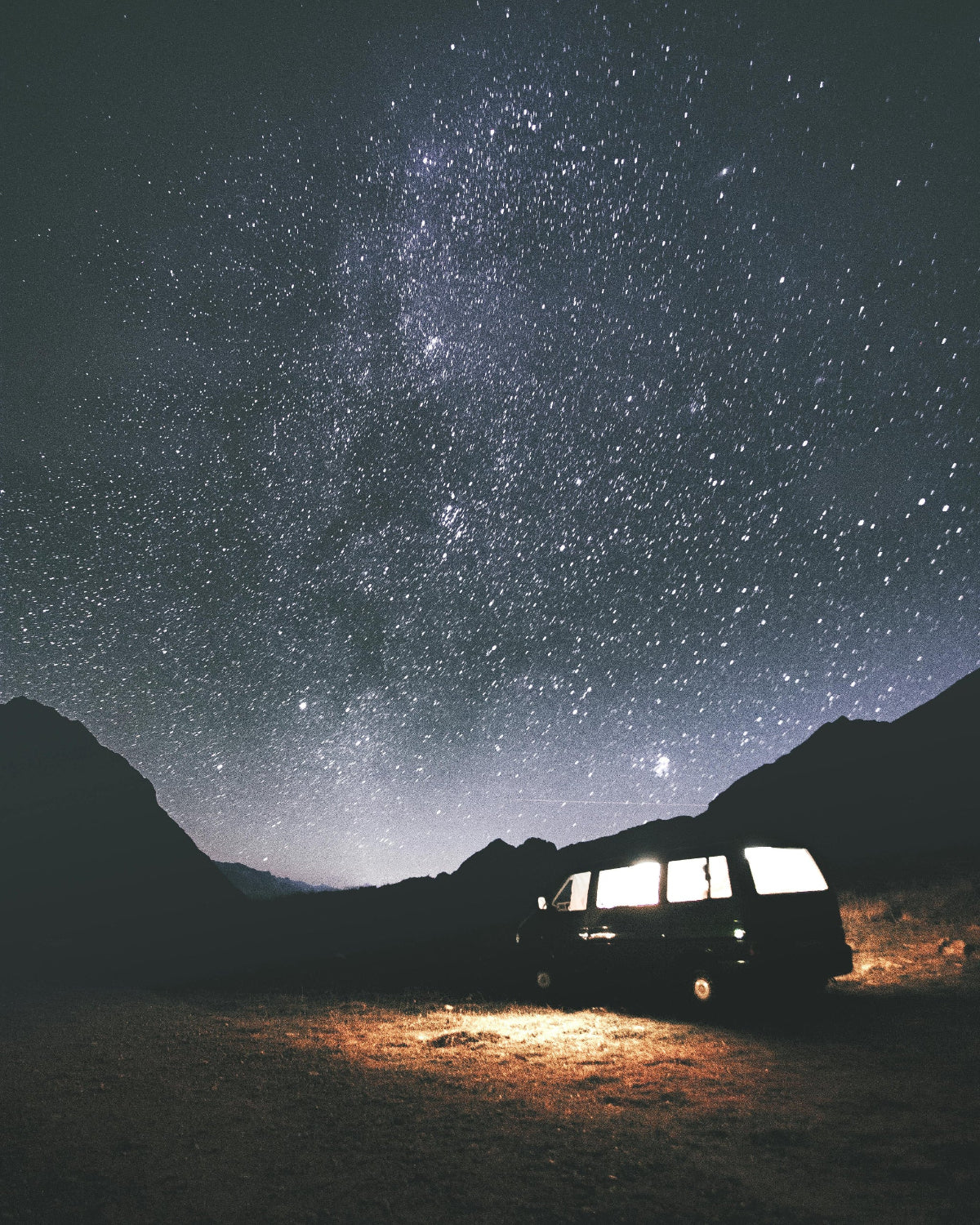 Camper van illuminated with HAVEN solar portable lantern and power bank at mountainous campsite overlooking dark starry sky.