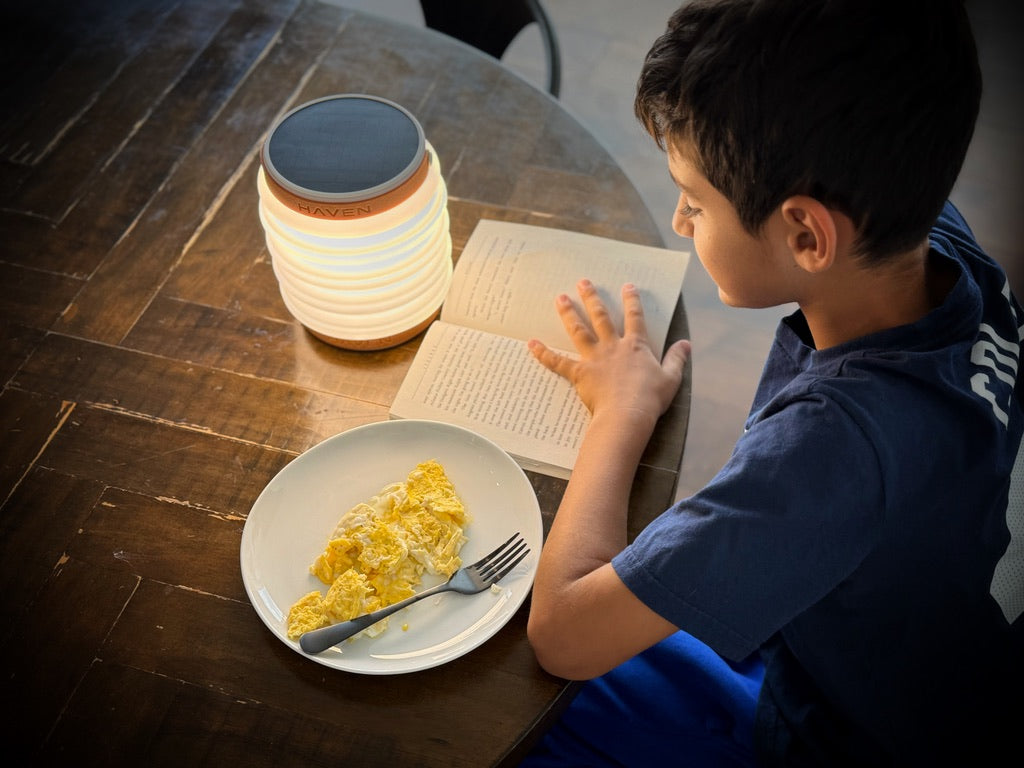 Child reading a book using a HAVEN solar portable lantern while sitting at a dining room table for breakfast.  