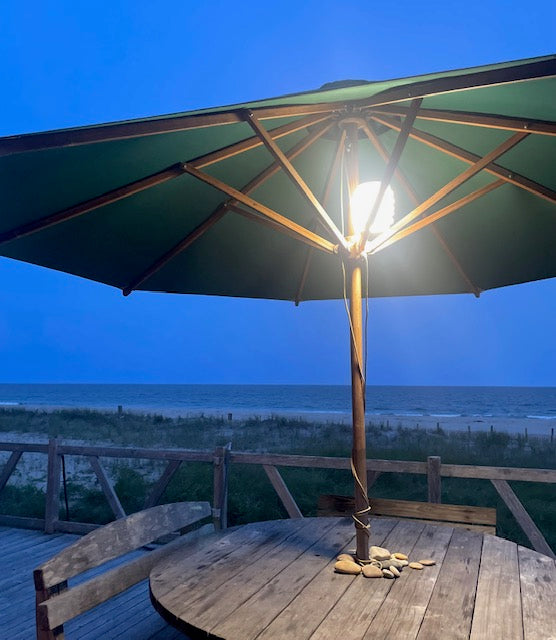 HAVEN portable solar lantern hanging from a patio umbrella on a wooden table with a beach view in the background.