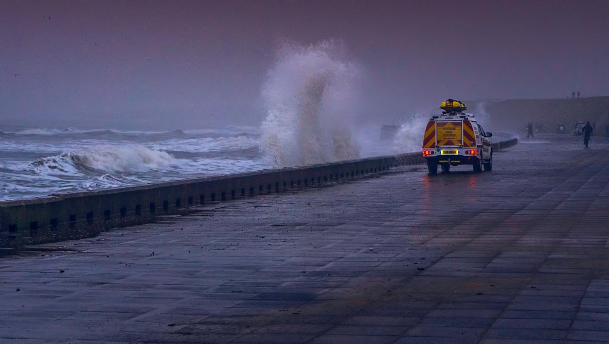 Emergency vehicle on a coastal road during a storm with large waves crashing against the wall.