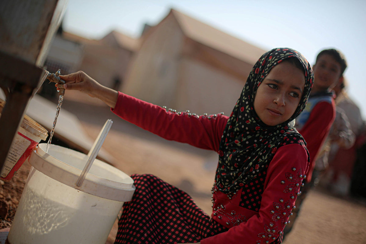 Young woman in a patterned headscarf filling a container with water in a refugee camp.